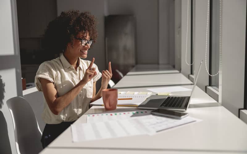 Mujer sonriente frente a una computadora portátil, realizando una videollamada mientras se comunica en lengua de señas. Está sentada en un espacio de oficina iluminado con luz natural.