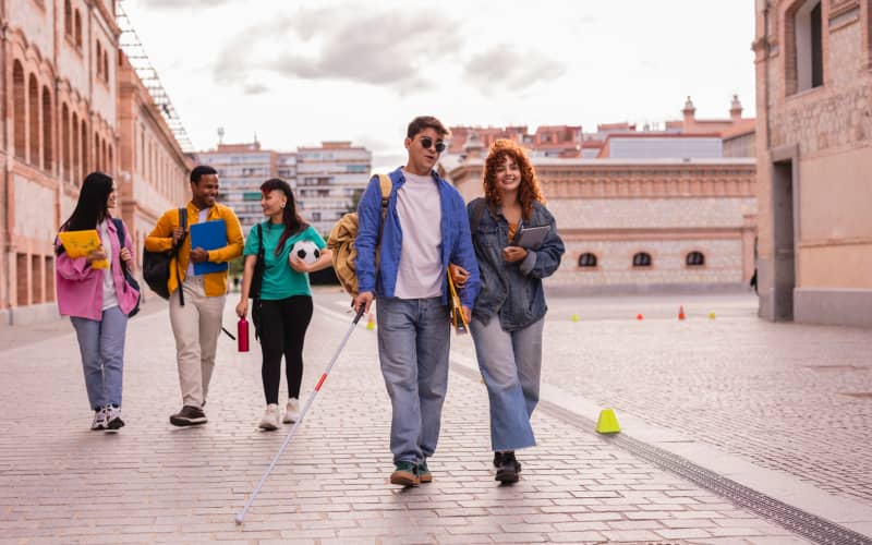 diversidad-funcional-discapacidad-2 Grupo de jóvenes caminando por una calle: uno de ellos, con bastón blanco y gafas oscuras, en representación de las dudas entre diversidad funcional o discapacidad visual.