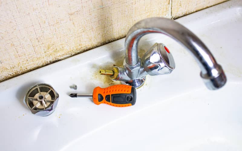 Un grifo bimando de lo que parece un baño con herramientas de bricolaje y algunas piezas alrededor, en representación e la acción de arreglar un grifo que gotea.