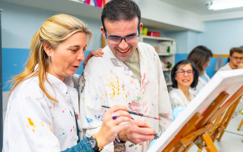 Persona con discapacidad participando en una clase de pintura guiada por una instructora, representando la inclusión social y los beneficios fiscales por discapacidad del 65 por ciento que apoyan programas de integración
