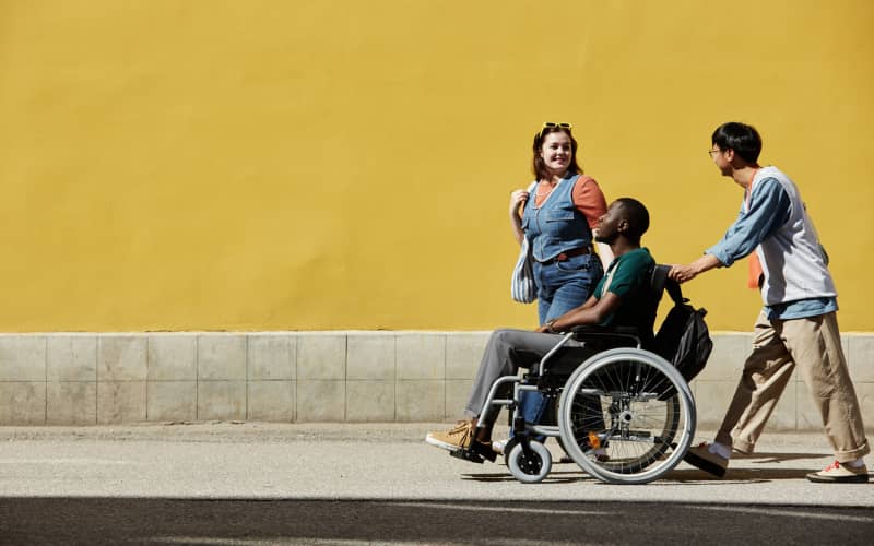 Tres personas caminan por la calle y una de ellas se desplaza en silla de ruedas, acompañada por sus amigos, ilustrando una situación de discapacidad sobrevenida y apoyo social