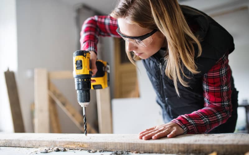 Chica joven utilizando herramientas en lo que parece un taller de bricolaje y demostrando como usar un taladro para agujerear una madera.
