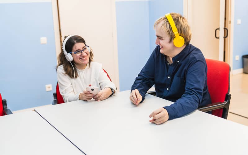 Dos personas con discapacidad sobrevenida sonríen mientras escuchan música con auriculares en una sala, compartiendo un momento de ocio y conexión