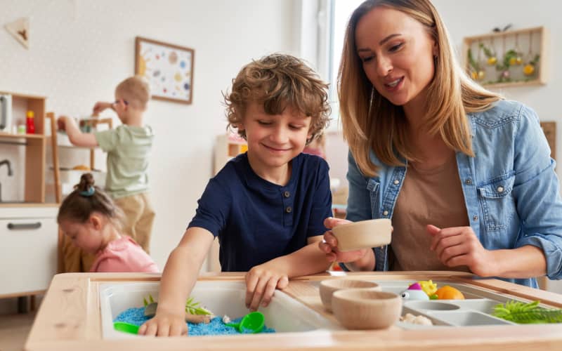 Educadora acompaña a un niño durante una actividad de juego con arena y objetos táctiles en un aula infantil, fomentando el aprendizaje y la inclusión de niños con discapacidad sensorial