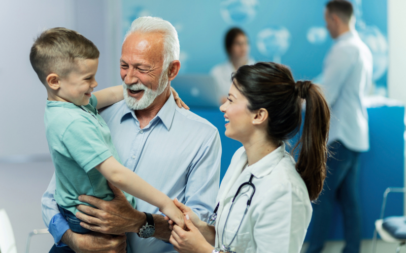 Abuelo con su nieto en brazos saludando a una doctora en una clínica, representando diferentes edades cubiertas por un seguro de salud.