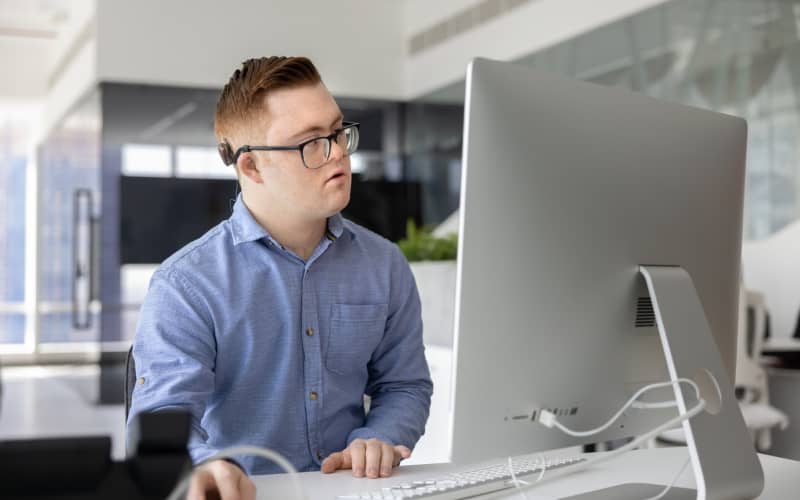 Joven con discapacidad intelectual trabajando concentrado frente a un ordenador de sobremesa en una oficina moderna, contribuyendo a cubrir esa cuota de discapacidad en empresas.