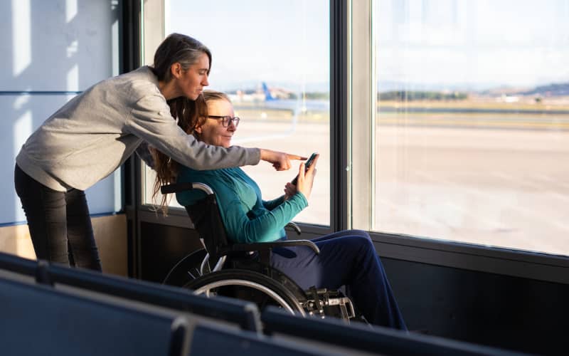 Mujer acompañando a otra persona en silla de ruedas mientras consultan información en un teléfono móvil junto a un ventanal, en representación de situaciones de lectura fácil.