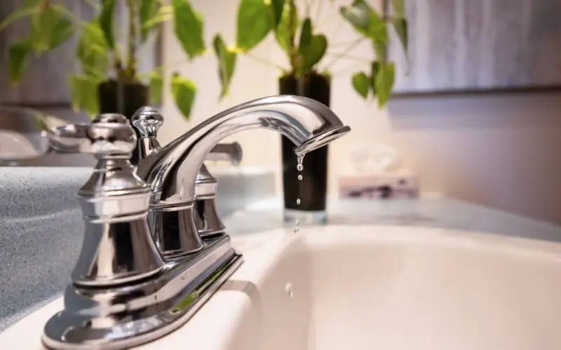 Primer plano de un grifo bimando colocado en el lavabo de un baño y que está goteando, en representación de la necesidad de arreglar un grifo que gotea.