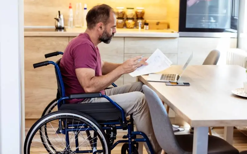 Una persona en silla de ruedas revisa documentos en la mesa de la cocina de casa, en representación de los beneficios fiscales por discapacidad del 65%
