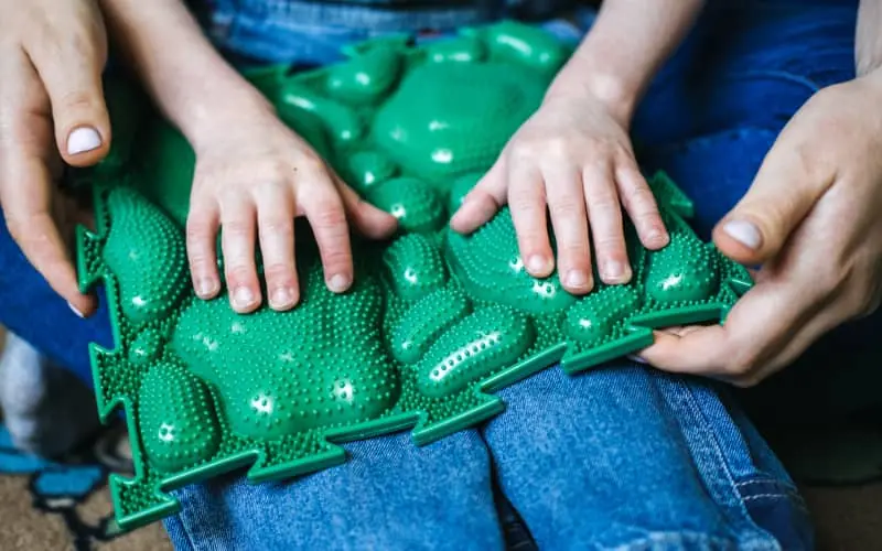 Manos de un niño y un adulto explorando un panel táctil verde con relieves, material de estimulación y apoyo para la discapacidad sensorial