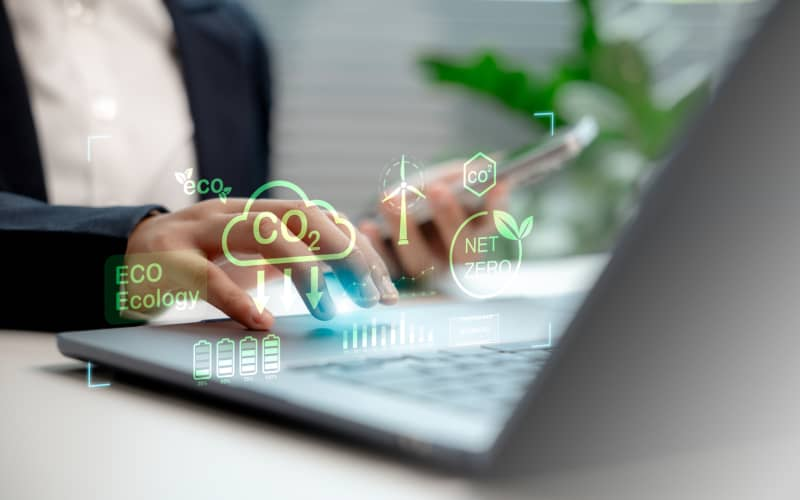 Manos de una persona escribiendo en su ordenador portátil con un diseño gráfico de emisiones sobreimpresos, en representación de la huella de carbono personal.