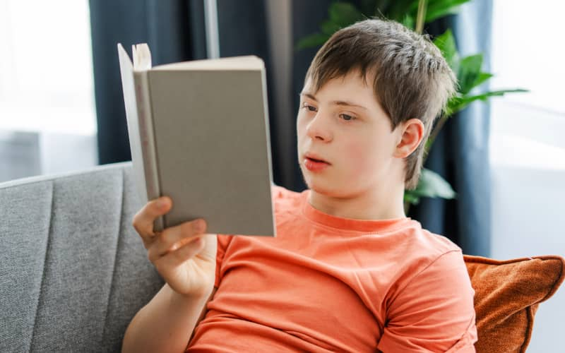 Niño con Síndrome de Down sentado en el sofá de una casa con un libro en la mano y leyendo, en representación de una situación de lectura fácil.