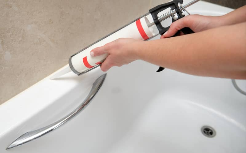 Unas manos colocando silicona para sellas las juntas de una bañera con una pistola de silicona en un baño limpio.