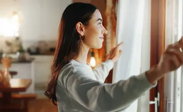 Mujer abriendo las cortinas de una ventana, preparando el espacio para ventilar antes de aplicar soluciones sobre cómo aislar ventanas y mejorar la eficiencia energética.