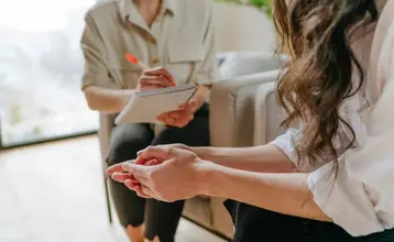 discapacidades_-invisibles-00.jpg Mujer en sesión de terapia psicológica con una profesional de la salud mental, ambas sentadas y conversando en un entorno cómodo. La imagen representa el apoyo profesional en casos de discapacidad invisible.