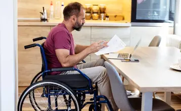 Una persona en silla de ruedas revisa documentos en la mesa de la cocina de casa, en representación de los beneficios fiscales por discapacidad del 65%