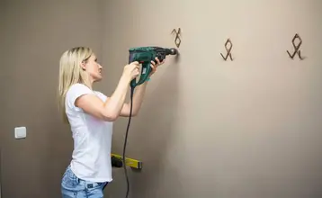 Una mujer joven y fuerte vestida con vaqueros y camiseta básica blanca de manga corta está haciendo agujeros en una pared marrón con un taladro eléctrico.