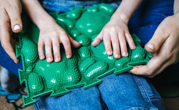 Manos de un niño y un adulto explorando un panel táctil verde con relieves, material de estimulación y apoyo para la discapacidad sensorial