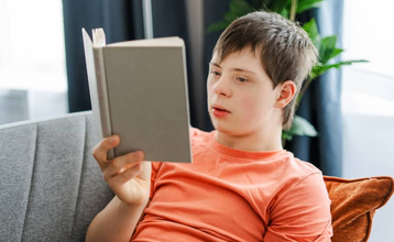 Niño con Síndrome de Down sentado en el sofá de una casa con un libro en la mano y leyendo, en representación de una situación de lectura fácil.