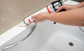 Unas manos colocando silicona para sellas las juntas de una bañera con una pistola de silicona en un baño limpio.